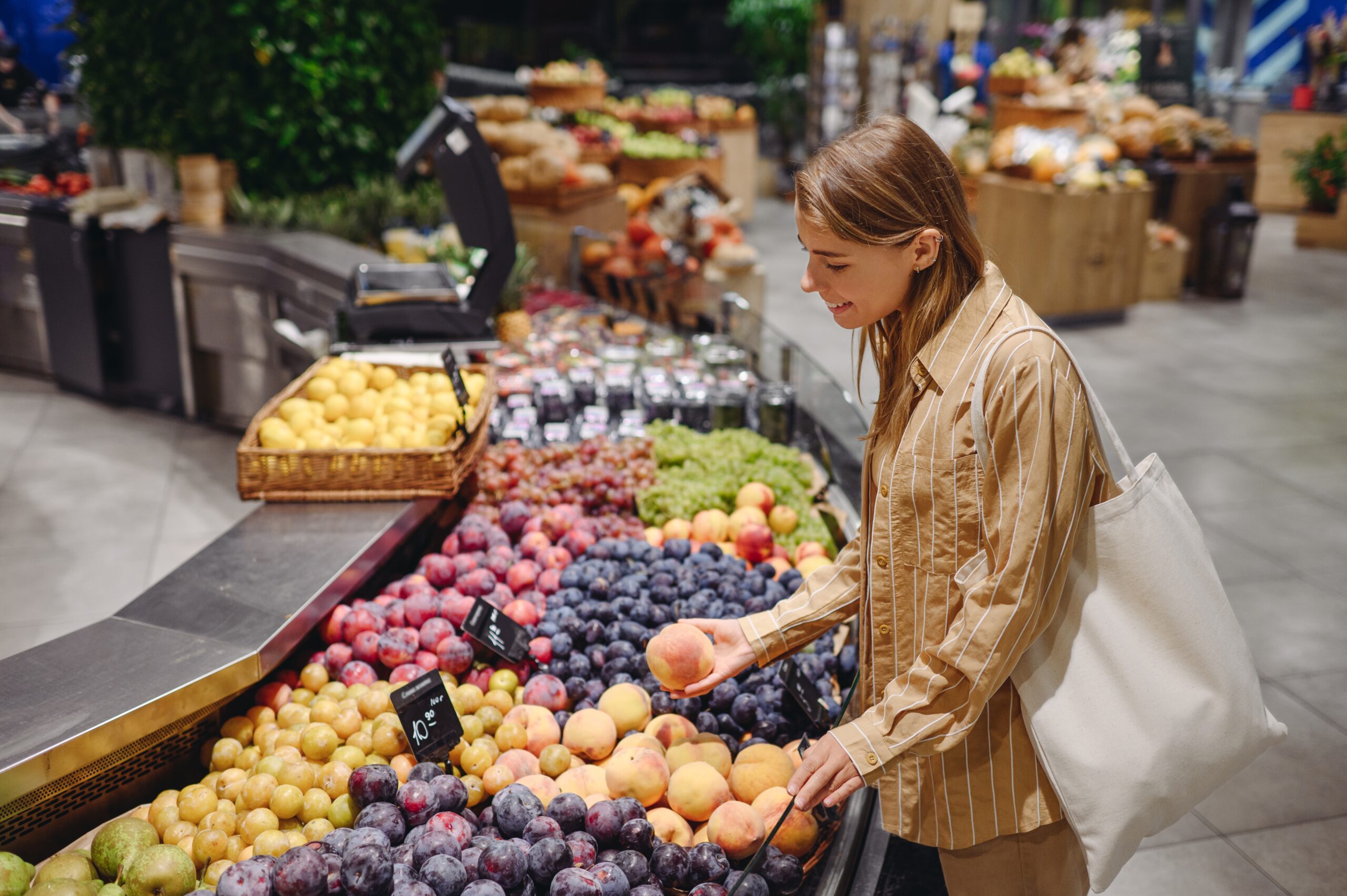 A woman smiles as she looks down and picks up a peach from a display of fruit in a supermarket