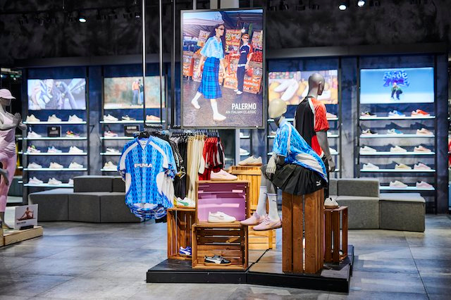 A Puma display in a store. A screen showing a woman in puma clothing is hung above mannequins wearing puma clothing and a rack of puma shirts.