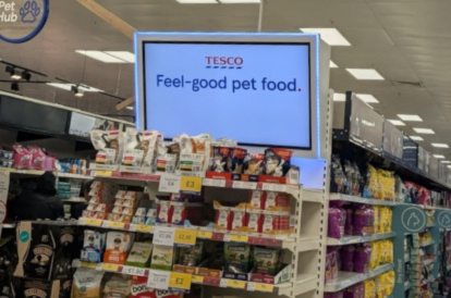 A photo of an interior of a Tesco store. The image shows the end of an aisle. On the top is a digital screen that says Feel Good Pet Food.