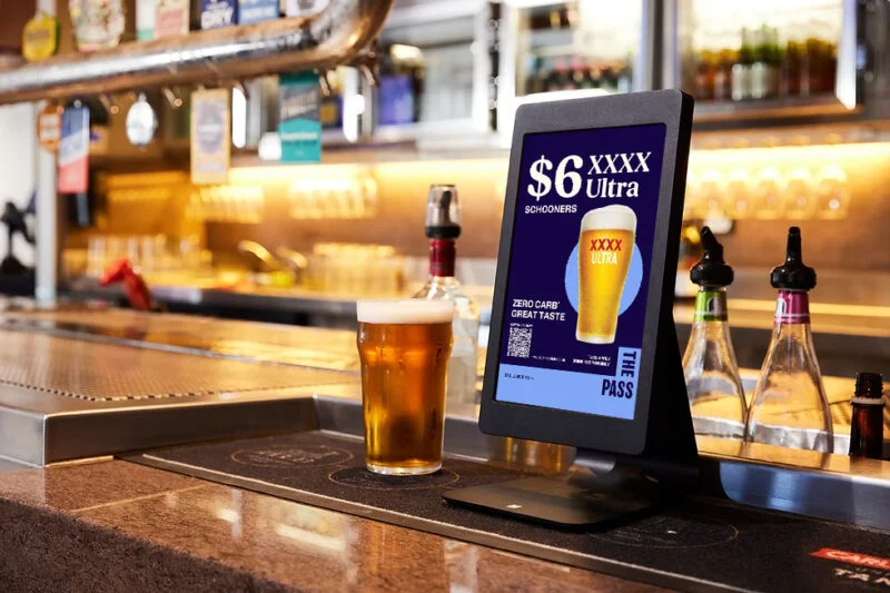 A photo of the bar within a pub. A pint of beer sits in front of vertical tablet showing a promotional advert for a beer