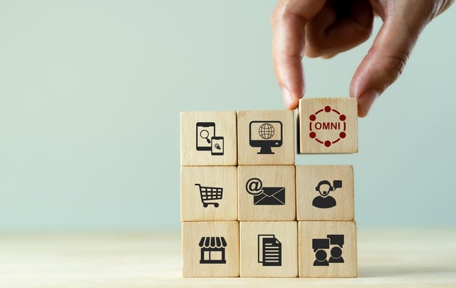 A photo of a pair of fingers placing a building block with the word omni on it on top of block which contain shopping related icons such as a shopping cart and a computer monitor.