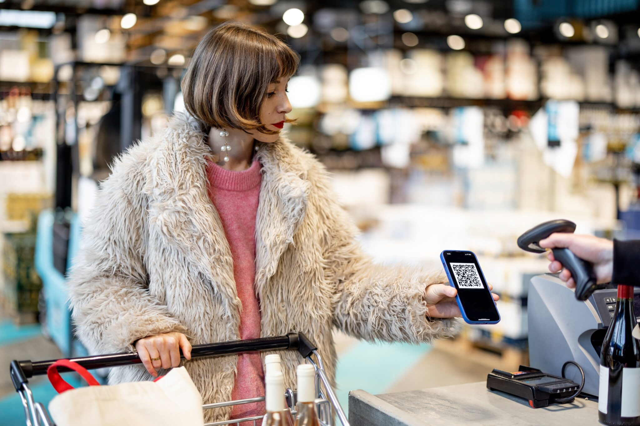 A photo of a stylishly dressed woman in a store. She is a pushing a shopping trolly and holding a cell phone to the register