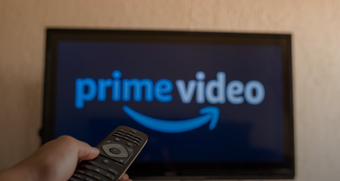 A photo of a hand extending a remote control towards a screen showing the Amazon prime video logo