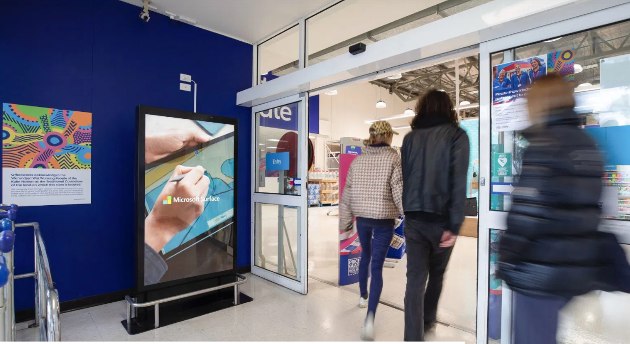 A photo of people entering what looks to be a large store passing a digital screen.
