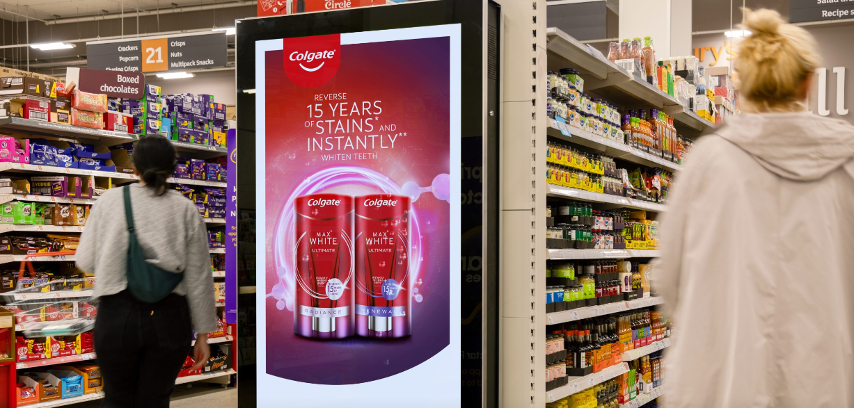 A photo of an interior of a supermarket. The asiles are filled with products and a digital; advert for toothpaste can be seen on the end of one of the aisles.
