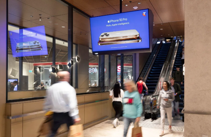 a photo of the inside of a shopping center. An escalator is visible as is an overhead digital screen on which a cell phone advert can be seen.