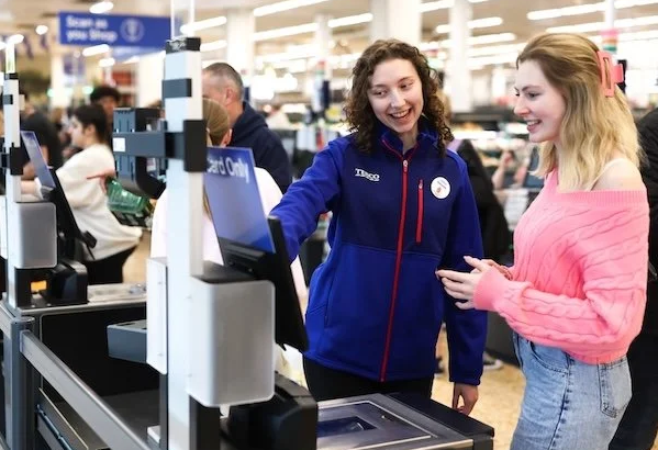 a photo at a self service check out in a supermarket. A member of staff smiles as she helps a customer.