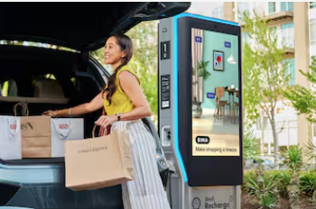 A photo of a woman putting parcels into the trunk of her car. She is smiling. Behind her a Volta charging station is visible.