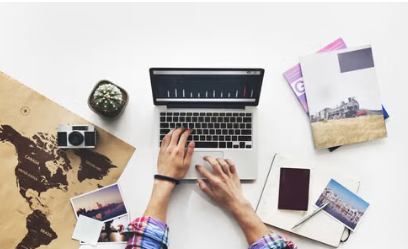 An overhead photo of hands on a laptop. Around the laptop is a passport, photos and maps - items that suggest travel.