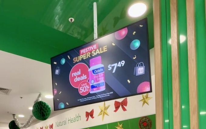 A photo of a landscape orientated screen hung from a ceiling. On the screen, a promotion for a product in a pink labelled bottle can be seen