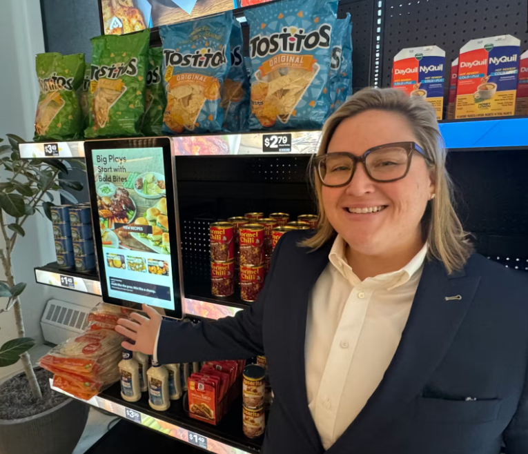 Liz Roche smiles while gesturing to a digital screen on a supermarket shelf.